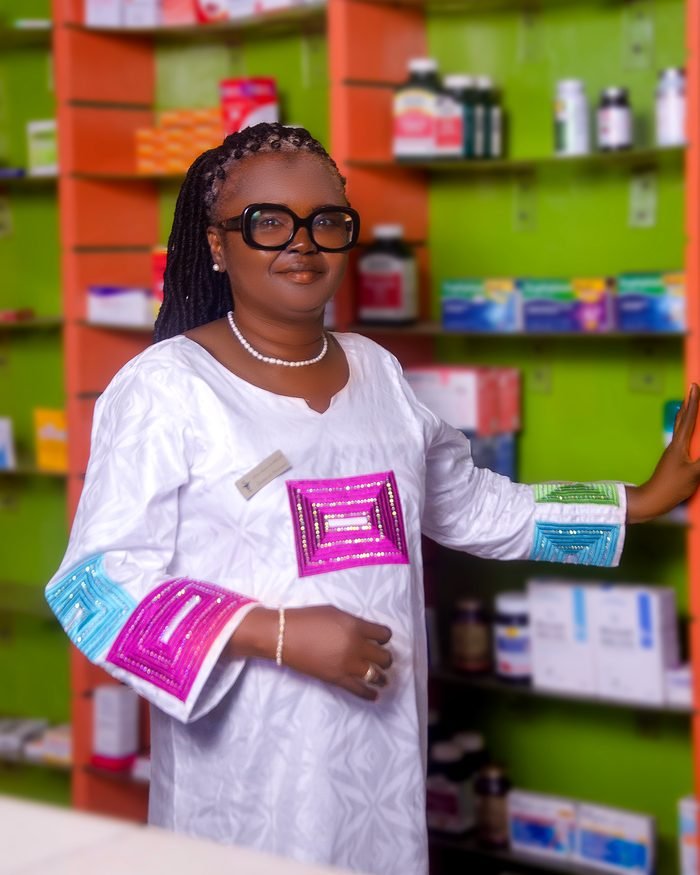 Dr Raïma MALICK debout dans l'officine de la Pharmacie de l'Étoile, entourée des rayonnages de médicaments et produits de santé